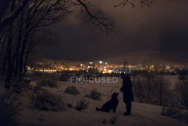 El hombre y su mascota caminan por la noche en el bosque nevado en un invierno cercano - foto de stock