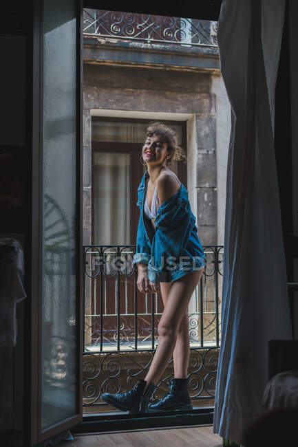 Junge, magere Frau in weißer Spitzenunterwäsche und Jeansjacke steht auf kleinem Balkon und blickt mit erhobener Hand in die Kamera — Stockfoto