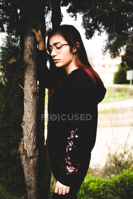 Jovem morena mulher em roupa preta e óculos de pé ao lado da árvore — Fotografia de Stock