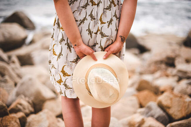 Vue arrière de la femme de la culture tenant chapeau et debout sur la côte rocheuse à l'océan — Photo de stock