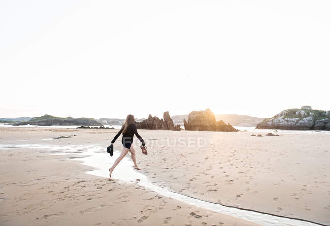 Barefoot woman holding hat and footwear while jumping over stream on sandy beach — Stock Photo