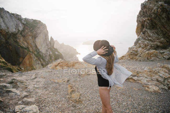 Woman in hat shirt standing on rocky coast and looking at view — Stock Photo