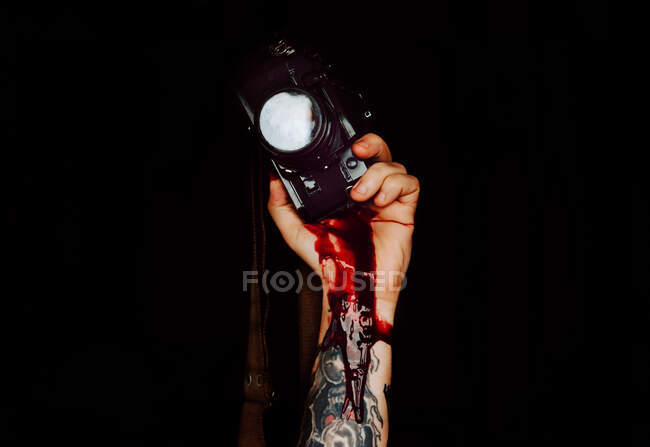 Recorte de fotografía de mano tatuada sosteniendo la cámara fotográfica con gruesa sangre oscura corriendo sobre fondo negro - foto de stock
