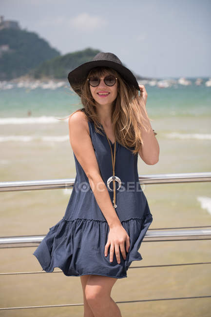 Young woman in hat and dress leaning on fence on background of sea — Stock Photo