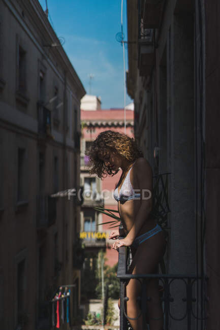 Vista lateral da jovem sedutora com cachos vestindo roupa interior de renda branca e em pé na varanda ensolarada — Fotografia de Stock