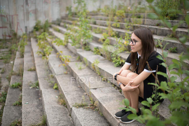 Vista lateral da jovem mulher em vestido preto e óculos sentados em passos cinza coberto e abraçando joelhos olhando para longe — Fotografia de Stock