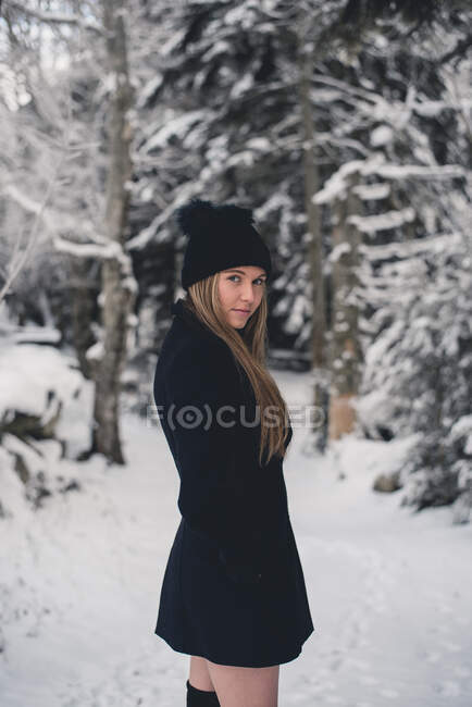 Jovem mulher atraente em chapéu engraçado e casaco preto posando no caminho da floresta e olhando para a câmera — Fotografia de Stock