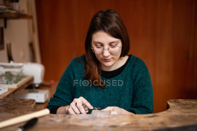 Konzentrierte Frau in grüner Bluse und Brille schnitzt Dekoration mit Instrument am Arbeitstisch in der Werkstatt — Stockfoto