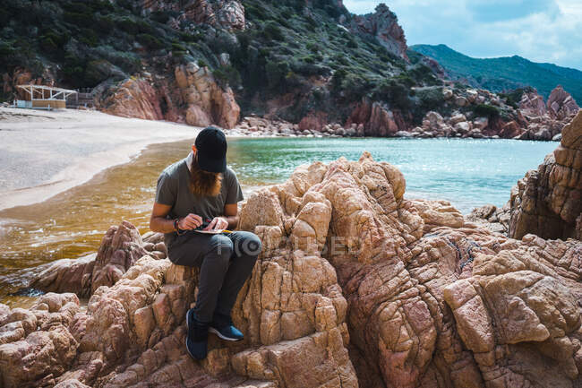 Uomo barbuto adulto seduto sulla pietra e prendere appunti al mare. — Foto stock