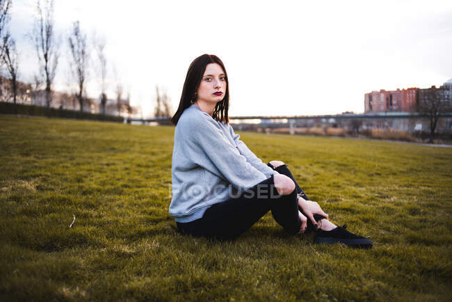 Seriöse Brünette in lässigem Outfit und dunklem Make-up sitzt auf grünem Gras und blickt in die Kamera. — Stockfoto