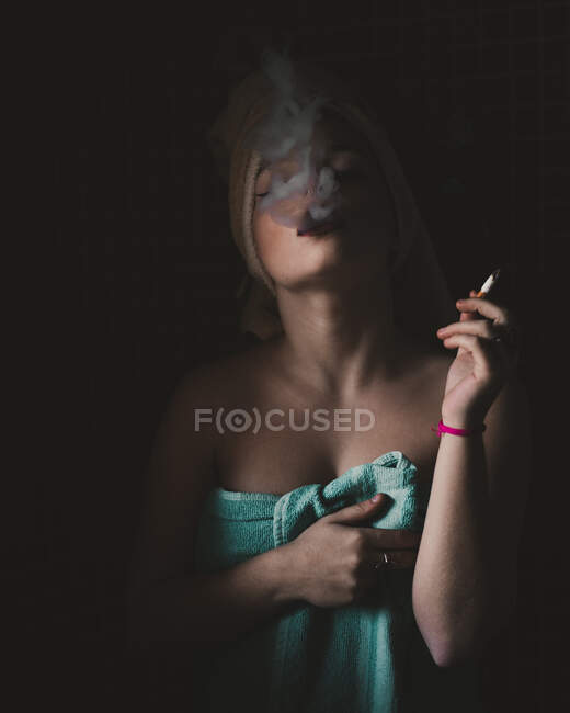 Jolie femme enveloppée dans une serviette debout et gonflant de la fumée sur fond sombre. — Photo de stock