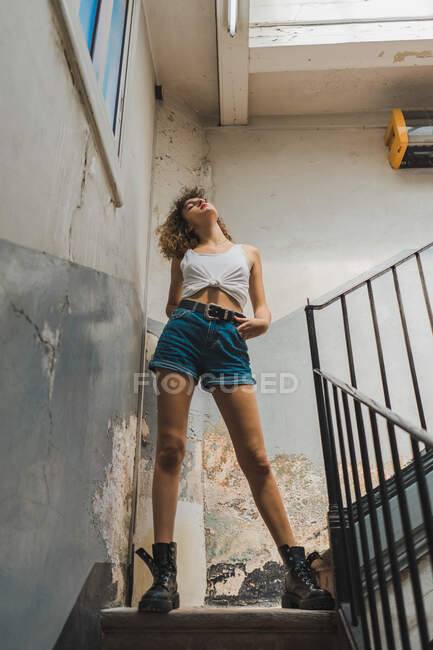Trendy giovane donna in pantaloncini e stivali con cappuccio in piedi sensualmente su scala con parete shabby — Foto stock