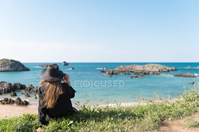 Frau in schwarzem Outfit und Hut sitzt auf Gras an der Küste und genießt den Blick auf das Meer — Stockfoto
