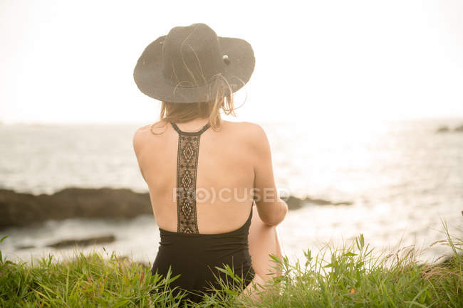 Woman in hat and swimwear sitting on grass on coast and looking at view — Stock Photo