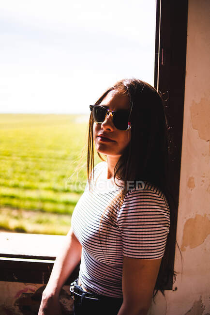 Jeune femme en lunettes de soleil debout près de la fenêtre et regardant beau champ vert par une journée ensoleillée. — Photo de stock