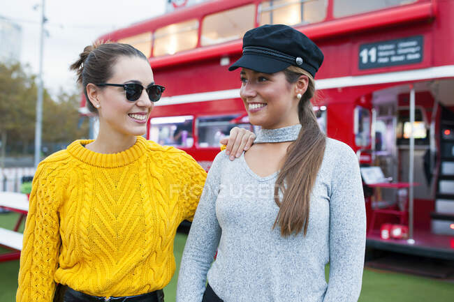 Zwei junge fröhliche Frauen in Pullovern und Accessoires stehen zusammen auf der Straße und schauen sich um. — Stockfoto