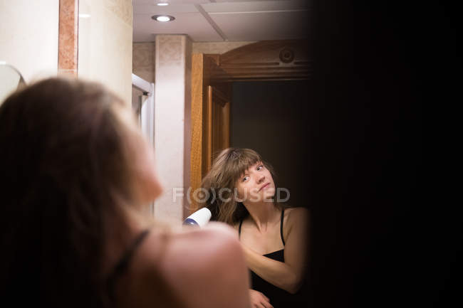 Молода жінка дивиться на дзеркало в стильній кімнаті і використовує сушарку для сушіння волосся — стокове фото
