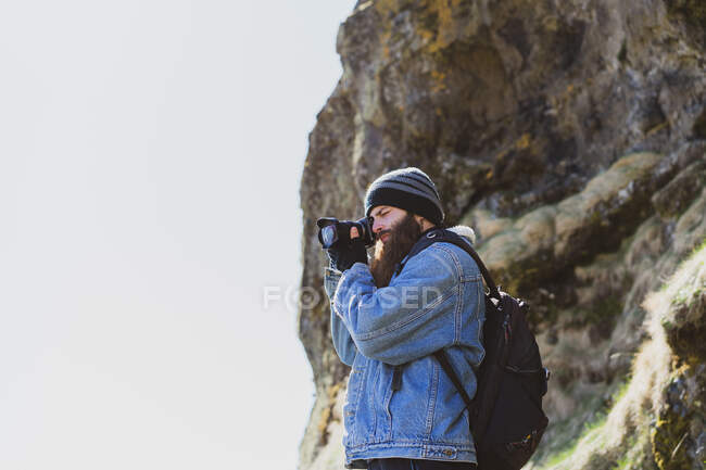 Боковой вид бородатого мужчины в шляпе с рюкзаком, снимающего на камеру исландский пейзаж — стоковое фото
