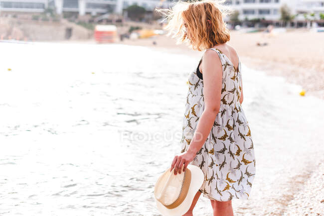 Visão traseira da mulher bonita segurando chapéu e em pé no oceano calmo na praia na baía — Fotografia de Stock