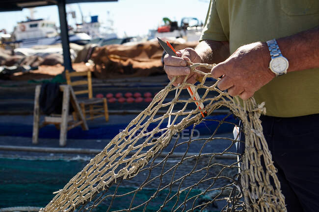 Crop from above view of man holding fishing net in hands and cutting with knife — Stock Photo