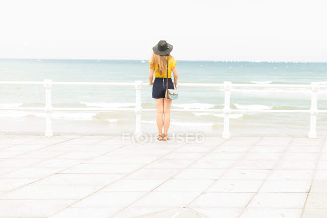 Woman in elegant outfit standing near fence on embankment and enjoying view of sea — Stock Photo