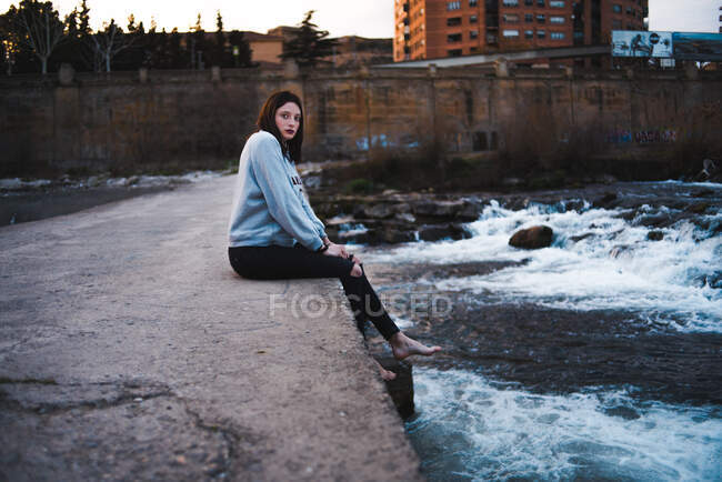 Side view of casual brunette in hoodie sitting barefoot on concrete waterfront with waves below and looking away. — Stock Photo