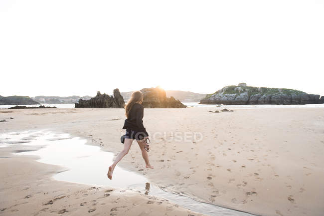 Donna scalza che tiene cappello e calzature mentre salta sopra il flusso sulla spiaggia sabbiosa — Foto stock