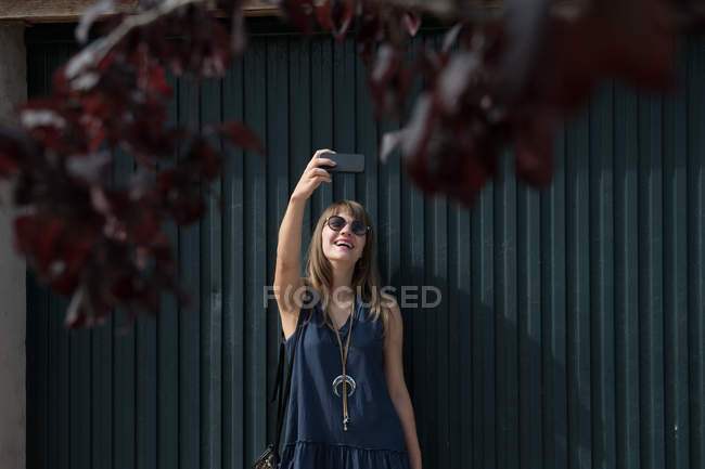 Joven mujer sonriendo y posando para selfie mientras de pie cerca de valla - foto de stock