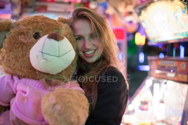 Cheerful woman embracing huge teddy bear gained in playing amusement machine and smiling at camera — Stock Photo