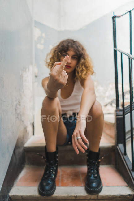 Young curly woman in brutal boots and shorts sitting on shabby stairway and showing middle fingers — Stock Photo