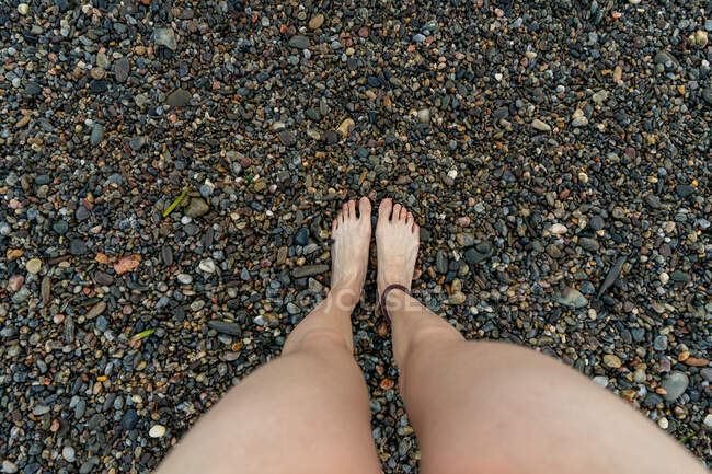De arriba plano de las piernas de la persona anónima de pie sobre pequeños guijarros en la costa pedregosa en Tavallera - foto de stock