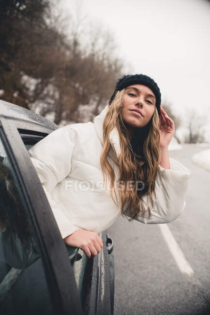 Mujer joven con el pelo rizado y sombrero divertido sonriendo de negro auto y mirando a la cámara - foto de stock
