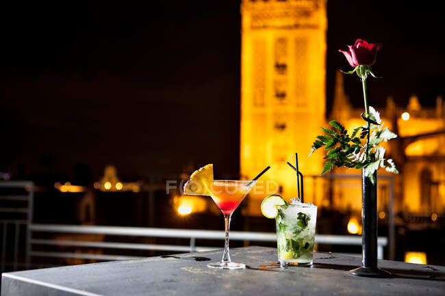 Glasses with cocktails and red rose in vase placed on table on roof on background of building lit by lantern at night — Stock Photo
