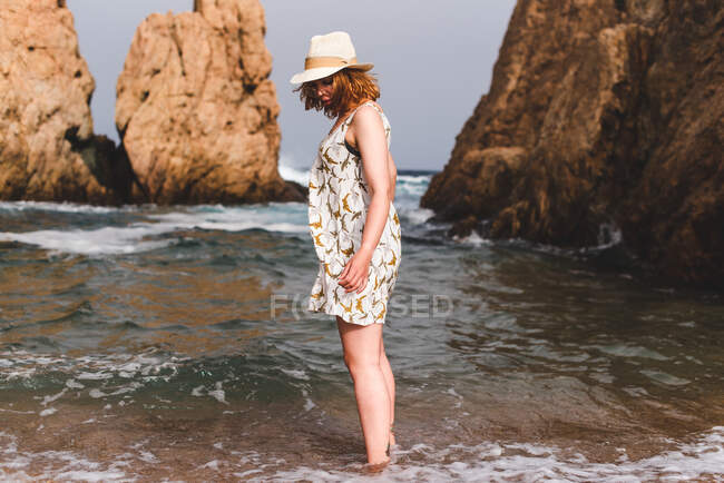 Pretty woman in hat standing and relaxing in the ocean at big rocks — Stock Photo