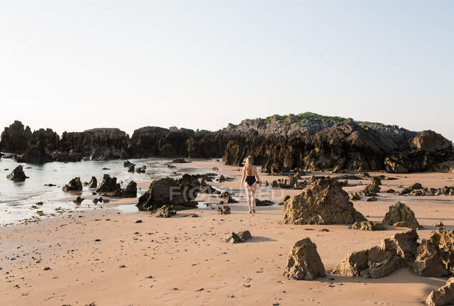 Silhouette di donna in costume da bagno a piedi sulla spiaggia di sabbia con rocce — Foto stock