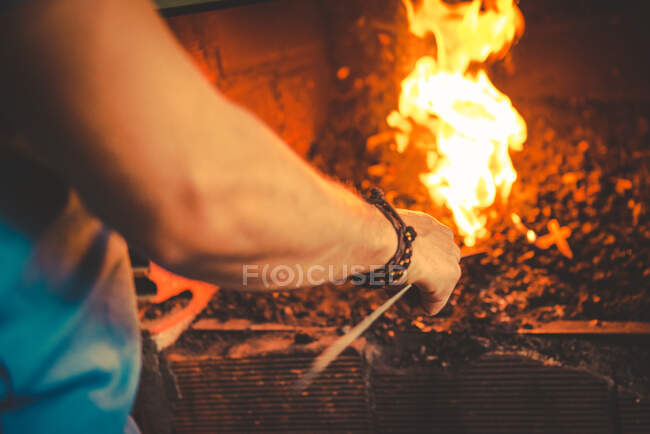 Ferreiro irreconhecível em pé perto do forno em chamas na oficina e peça de aquecimento de metal para forjar — Fotografia de Stock