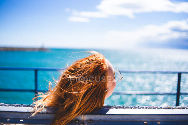 Vue arrière de jolie rousse assise et relaxante sur le banc à l'océan bleu par temps ensoleillé. — Photo de stock