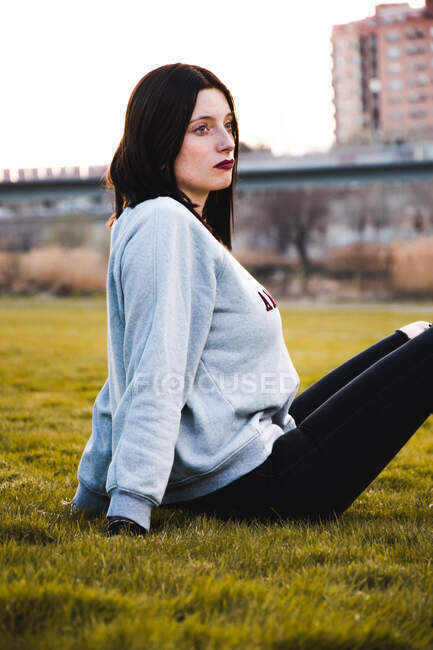 Brune sérieuse en tenue décontractée et avec un maquillage sombre assis sur de l'herbe verte regardant la caméra. — Photo de stock