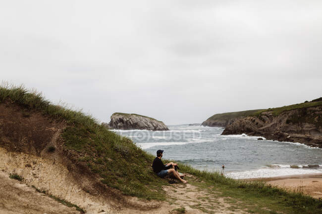Male sitting on grass on coast of sea and looking at water in Cantabria, Spain — Stock Photo
