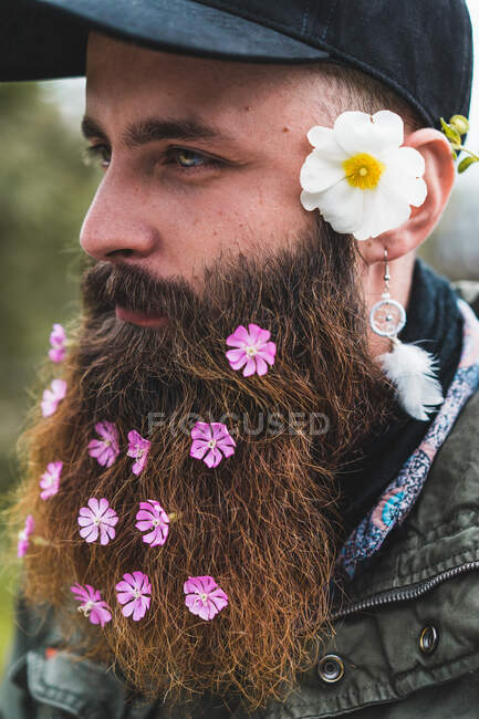Веселый взрослый мужчина с мягкими цветами в бороде делает селфи со смартфоном в природе. — стоковое фото