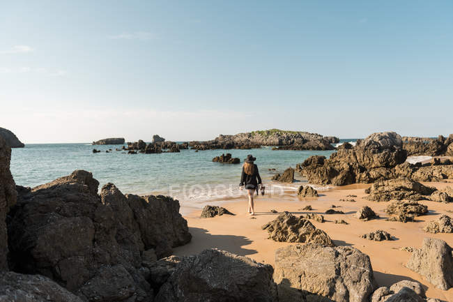 Mulher de chapéu andando na costa arenosa em meio a pedregulhos em direção ao mar — Fotografia de Stock
