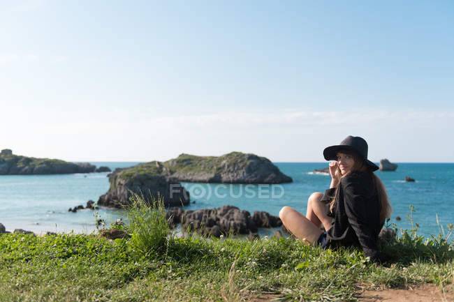 Женщина в элегантном наряде сидит на траве на берегу с видом на море и смотрит в камеру — стоковое фото