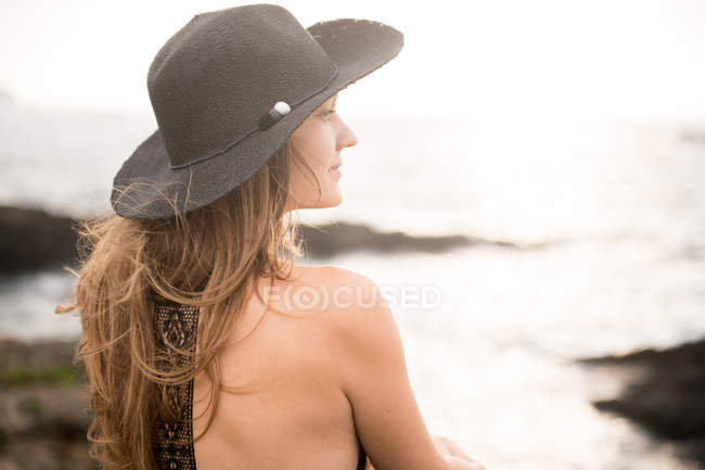 Primer plano de mujer joven en sombrero sentado en la costa y mirando a la vista - foto de stock