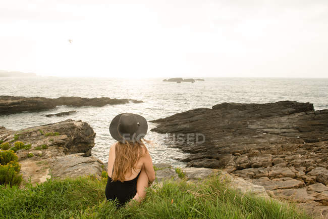 Femme en chapeau et maillots de bain assis sur l'herbe sur la côte et regardant la vue — Photo de stock