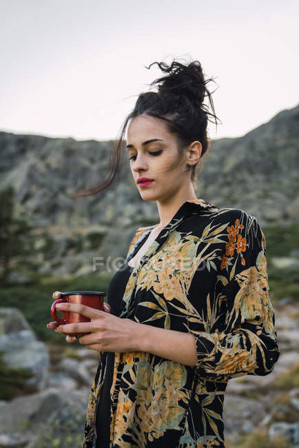 Giovane bella donna in piedi con tazza di caffè su pietre in campagna — Foto stock