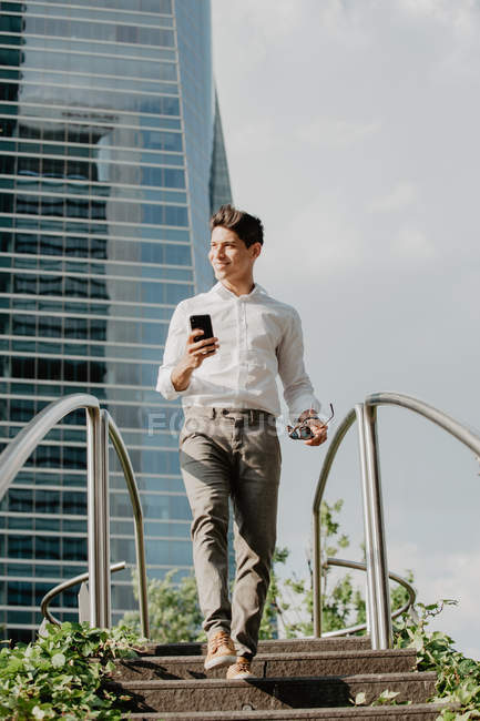 Cheerful man with smartphone walking down steps in modern city — Stock Photo