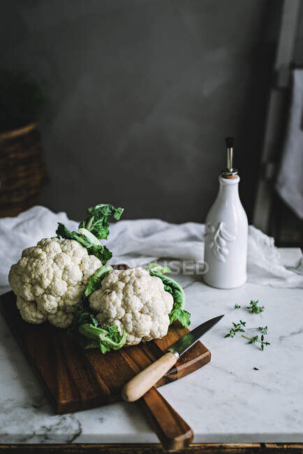 Cauliflowers com folhas verdes deitado em tábua de corte de madeira com faca sobre mesa de mármore com ervas e vaso com material têxtil sobre fundo quarto cinza em foco macio — Fotografia de Stock