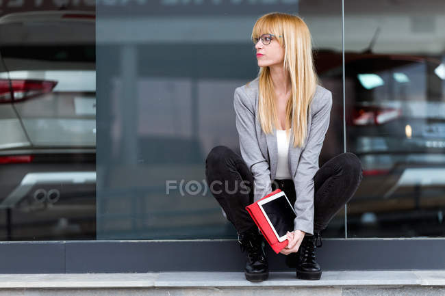 Молода приваблива жінка сидить з планшетом перед сучасною будівлею — стокове фото