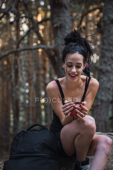 Young slim brunette holding metal mug and sitting on ground in woods with backpack — Stock Photo