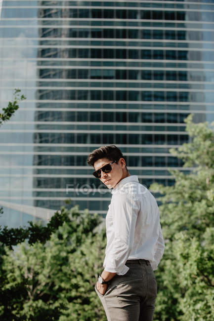 Jeune homme d'affaires en lunettes de soleil debout devant un bâtiment moderne et regardant par-dessus l'épaule — Photo de stock